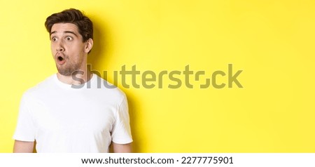 Close-up of impressed man looking left, gasping amazed, standing in white t-shirt against yellow background. Royalty-Free Stock Photo #2277775901