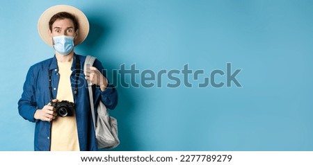 Covid-19 and summer holidays concept. Excited tourist in straw hat and medical mask looking in awe, holding camera, taking pictures on holiday during pandemic, blue background.