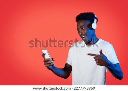 Joyful happy young black guy listening to music or watching vieo content online, using smartphone and wireless headphones, pointing at cell phone in his hand, red studio background, copy space Royalty-Free Stock Photo #2277793969