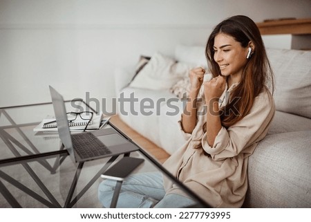 Cheerful millennial mixed race lady in glasses and wireless headphones has video call on laptop, make victory, success gesture in room interior. Great news, win in work, business remotely at home