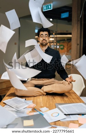 Vertical portrait of concentrated young business man sitting on table in lotus position with closed eyes, meditating despite of flying papers chaos surrounding at office, meditation and calmness. Royalty-Free Stock Photo #2277795661