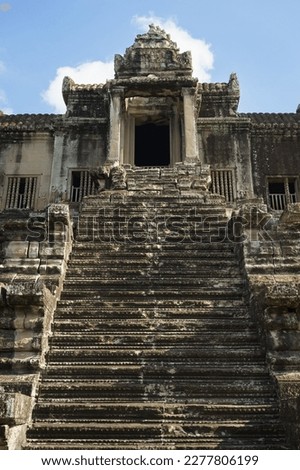 A photo looking up at one of the steep stairways leading to the upper tier of the temple mountain at Angkor Wat. Intricate carvings and traditional carved window balusters are visible. 