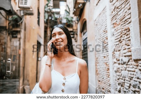 Happy Latin American woman looking away with smile and talking on smartphone near brick wall of aged building on narrow street of Barcelona, Spain Royalty-Free Stock Photo #2277820397