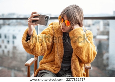 Beautiful young girl in a yellow fur coat is sitting in a director's chair on the terrace of a modern building. Using smartphone, social media influencer, taking picture, selfie, posing, city in back