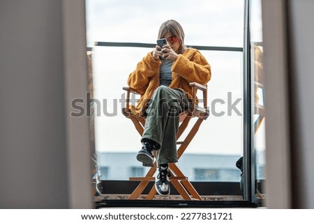 Beautiful young girl in a yellow fur coat is sitting in a director's chair on the terrace of a modern building. Using smartphone, social media influencer, taking picture, selfie, posing, city in back Royalty-Free Stock Photo #2277831721