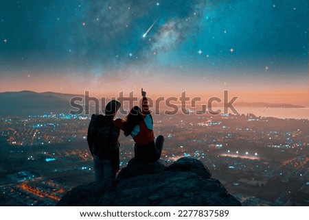 silhouettes of a couple sitting on the top of the mountain looking and pointing out at shooting star and milky way over the city lights on the horizon	 Royalty-Free Stock Photo #2277837589