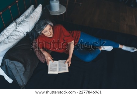 From above of middle aged female lying on bed with elbow support on pillow and looking away while reading book in hands in daylight and relaxing in bedroom at home Royalty-Free Stock Photo #2277846961