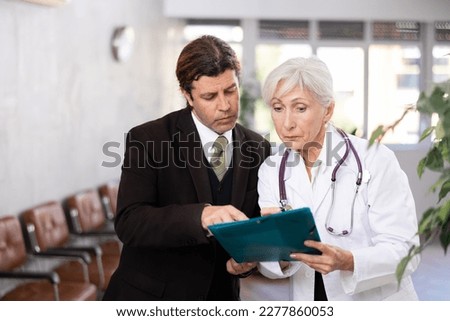 Adult man in business suit and elderly woman in medical uniform look at folder with documents in clinic reception..