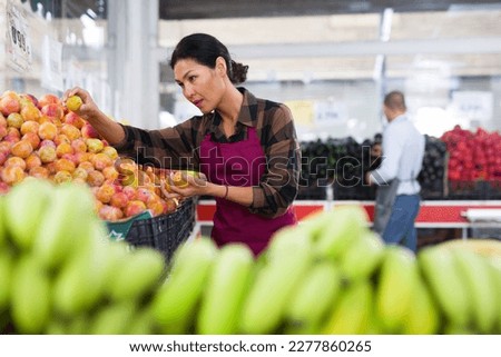 Asian woman in apron stacking plums in greengrocer. Man shopping in background.