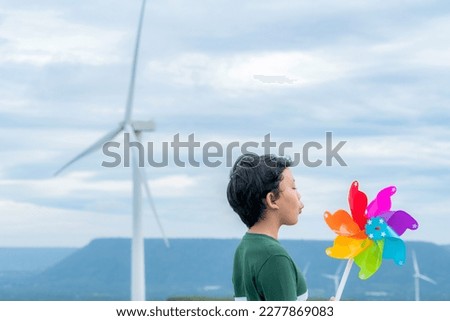 Progressive young asian boy playing with wind pinwheel toy in the wind turbine farm, green field over the hill. Green energy from renewable electric wind generator. Windmill in the countryside concept Royalty-Free Stock Photo #2277869083