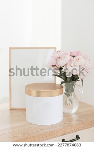 Vertical frame and gift box mockup on a wooden table in the kitchen. Glass jug with a bouquet of pink peonies. Scandinavian style interior.