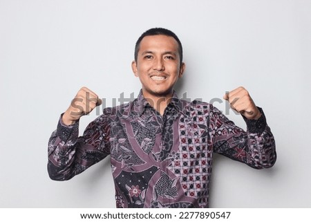 Asian man wearing a batik shirt showing strong gesture by lifting her arms and muscles smiling proudly on the white background.