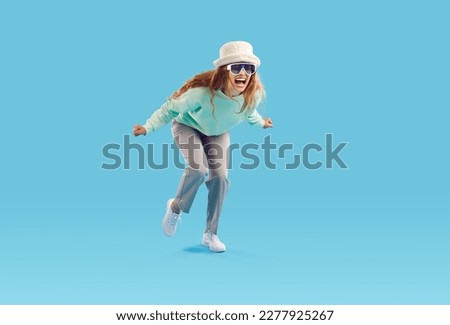 Studio shot of funny crazy screaming girl in fashionable outfit. Young woman in hat and sunglasses feeling happy and excited or furious and enraged screaming loudly isolated on blue background