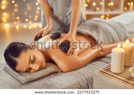 Woman getting exotic spa massage with hot stones. Happy, relaxed young woman lying on spa bed while professional masseuse is putting hot stones on her back. Spa treatment, body relaxation concept Royalty-Free Stock Photo #2277925279