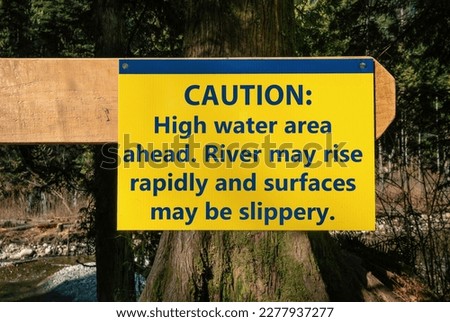 Trail caution sign about high water and fast rising river. Yellow large sign on fence pole in front of river from the park authority. Trail and hiking safety sign. Selective focus. 