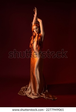 Full length fantasy portrait of beautiful woman model with red hair, goddess silk robes  gold crown. Standing pose gestural hands reaching out isolated on dark red studio background 