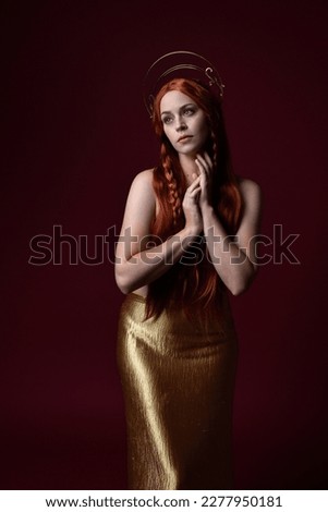 Close up fantasy portrait of beautiful woman model with red hair, goddess silk robes  gold crown.  Posing with gestural hands reaching out, isolated on dark red studio background 
