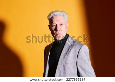 Serious mature businessman in grey blazer and black turtleneck looking at camera while posing against yellow background with shadows