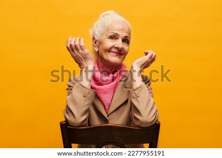 Senior skeptical woman sitting on chair with wooden back in front of camera in isolation and shrugging during photo session in studio