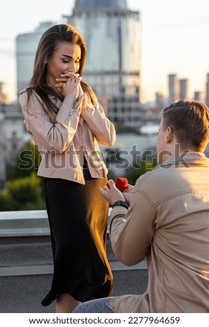 Handsome man is doing surprise marriage proposal to his beautiful girlfriend on rooftop standing on his knee. Happy loving couple on romantic date on Saint Valentine's Day. Urban cityscape SSTKHome