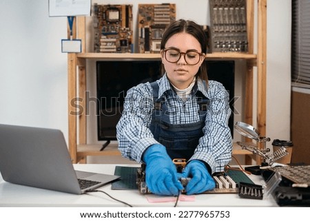 Tech fixes motherboard in service center. Female repairman working with computer with a part in hands. Monitors and other laptops in the background waiting for service. Copy space