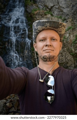Caucasian man wearing brown longsleeve shirt with sunglasses and khaki panama makes a selfie portrait with a small waterfall on the background. Hiker man makes photo of himself against the waterfall.