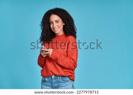 Young adult smiling happy pretty latin woman holding mobile phone device, doing ecommerce online shopping on cell, using apps on cellphone standing isolated on blue background. Royalty-Free Stock Photo #2277978715