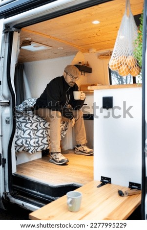 lonely man drinking coffee inside camper, thoughtful and serious person, van in the forest isolated and free. Camperized in cozy wood and grey and white colors. Comforter of gray hearts