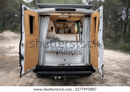 Back of camper with open doors, interior with wooden bed and kitchen, wooden doors, heart quilt and grey curtains, large white camper van, lonely forest interior. Royalty-Free Stock Photo #2277995887