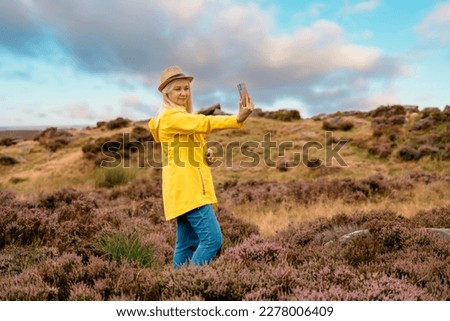 A woman in a yellow coat reaching the destination and  taking photo on top of a hill or mountain on sunny day in Peak Districkt.  Local tourism concept