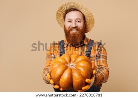 Happy smiling cheerful young bearded man wear straw hat overalls work in garden hold in hand give pumpkin isolated on plain pastel light beige color background studio portrait. Plant caring concept