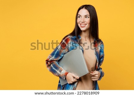 Side view young fun smart smiling IT woman wearing blue shirt beige t-shirt holding closed laptop pc computer look aside isolated on plain yellow background studio portrait. People lifestyle concept