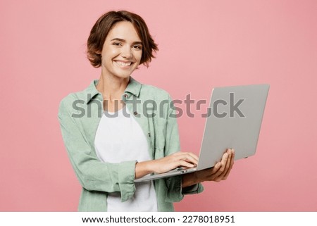 Young smiling happy cool IT woman 20s she wear green shirt white t-shirt hold use work on laptop pc computer isolated on plain pastel light pink background studio portrait. People lifestyle concept