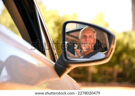 Senior Male Passenger With Mobile Phone Reflected In Wing Mirror Of Car Enjoying Day Trip Out