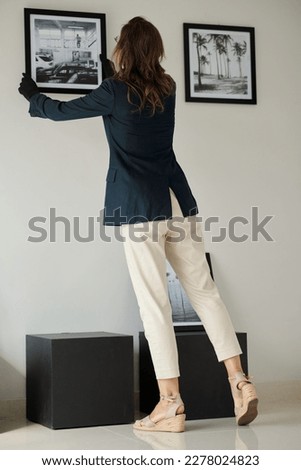 Rear view of young female in formalwear and black gloves hanging picture on wall