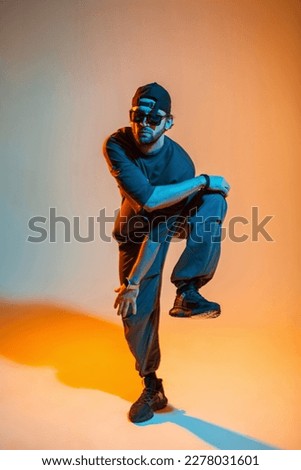 Stylish handsome professional dancer man with a cap and sunglasses in a trendy fashion t-shirt, pants and sneakers is dancing in a colorful studio with a creative orange and neon light