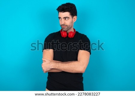 Pleased Young caucasian man wearing black T-shirt over blue background keeps hands crossed over chest looks happily aside