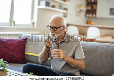 Senior man with glass of water taking pill at home. Age, medicine, healthcare and people concept. Mature man taking pill against headaches at home. Gray hair man at home taking pill to ease headache.