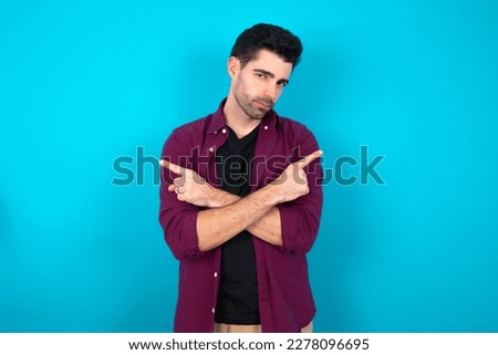 Serious Young man standing over blue studio background crosses hands and points at different sides hesitates between two items. Hard decision concept