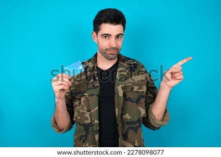 Smiling Young man standing over blue studio background showing debit card pointing finger empty space