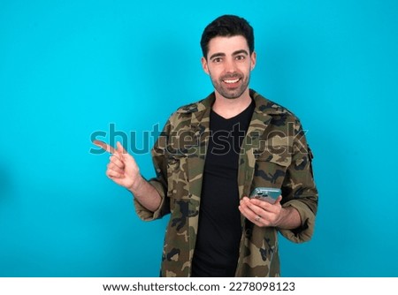 Astonished Young man standing over blue studio background holding her telephone and pointing with finger aside at empty copy space
