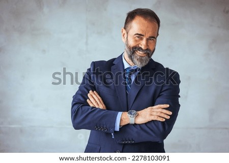 Happy mature business professional standing with his arms crossed in modern office. Smiling businessman at his workplace. Successful Mature man with folded arms standing over grey background. 
