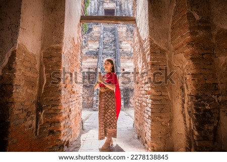 young woman dressed in traditional red Thai dress and golden ornaments stands sawasdee as a welcome and greeting in the ancient site at Wat Chaiwatthanaram, Ayutthaya, Thailand