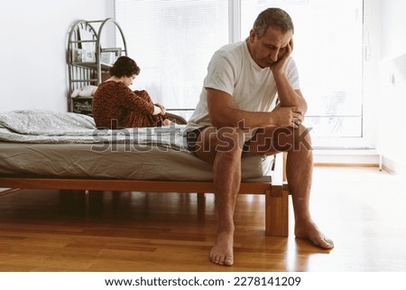 middle-aged man sits on edge of bed in a deplorable state, propping head in hands, young woman sits on opposite edge. Quarrel in bedroom Royalty-Free Stock Photo #2278141209