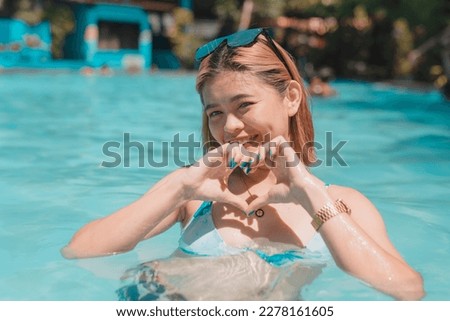 A cheerful young woman relaxing in chest deep waters in the swimming pool. Making a heart shaped sign with her hands. Enjoying her summer vacation.