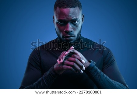 Determined adult bearded African American Athlete in black sportive turtleneck standing with folded arms in dark room with neon light and looking at camera Royalty-Free Stock Photo #2278180881