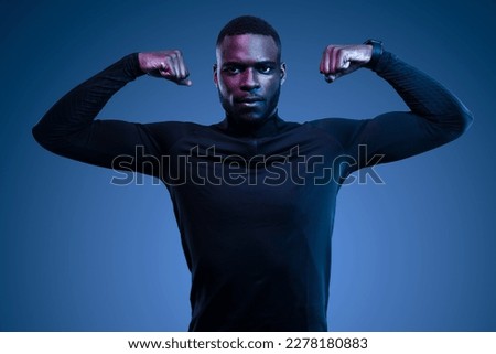 Self assured young bearded African American male athlete in stylish black activewear showing biceps and looking at camera against blue background Royalty-Free Stock Photo #2278180883