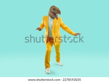 Cool man wearing rubber dinosaur head mask with open mouth. Stylish creative person dressed trendy yellow suit standing in crouching pose over light blue studio background