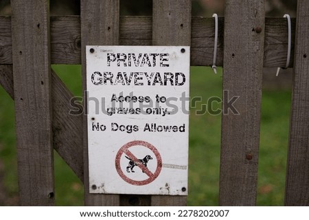 White "Private graveyard Access to graves only. No Dogs Allowed" sign with a red and black no dog symbol nailed screwed to wooden fence in front of grass of a cemetery in London, England.