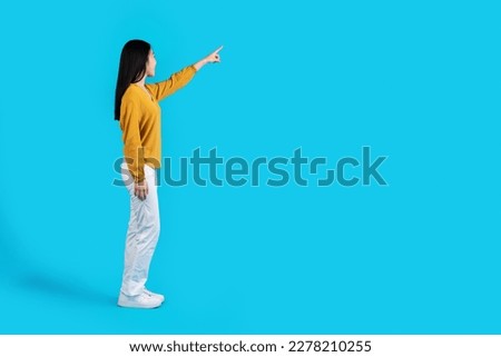 Side view of slim beautiful long-haired young asian woman in casual outfit pointing at free space for advertisement or text, isolated on blue studio background, full length shot Royalty-Free Stock Photo #2278210255